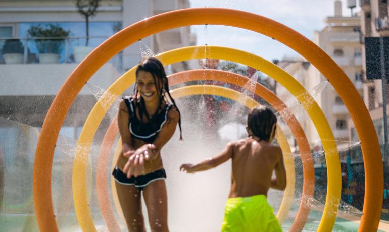 Bambini giocano sotto gli spruzzi d'acqua in un'area giochi all'aperto.