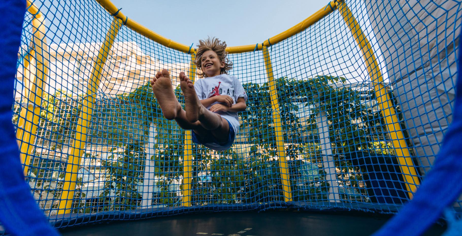 Bambino felice salta su un trampolino circondato da rete di sicurezza blu e gialla.