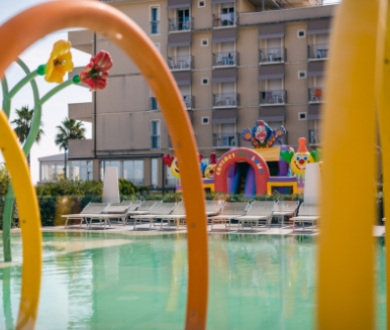 Piscina con giochi d'acqua e gonfiabili colorati davanti a un hotel.