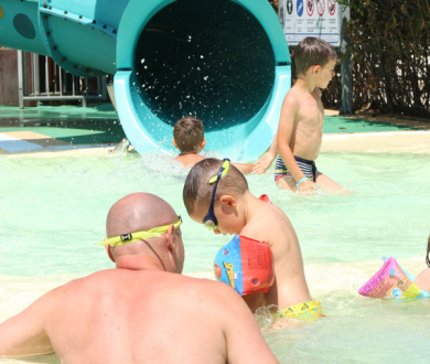 Bambini e adulti giocano in piscina con uno scivolo acquatico.