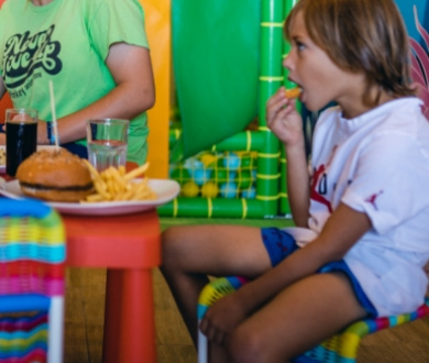 Bambino mangia patatine fritte accanto a un hamburger in un ambiente colorato.