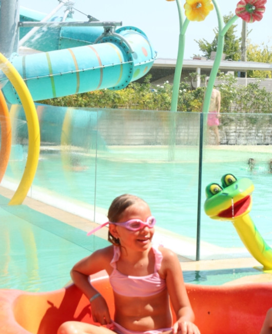 Bambina in costume rosa, occhialini, in piscina con scivolo e decorazioni colorate.