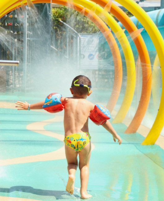 Bambino con braccioli gioca sotto spruzzi d'acqua in un parco acquatico.