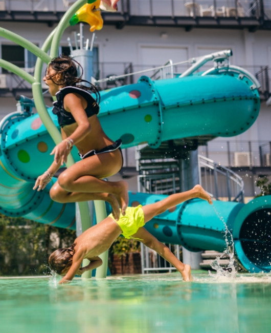 Due persone si divertono saltando in piscina con uno scivolo sullo sfondo.